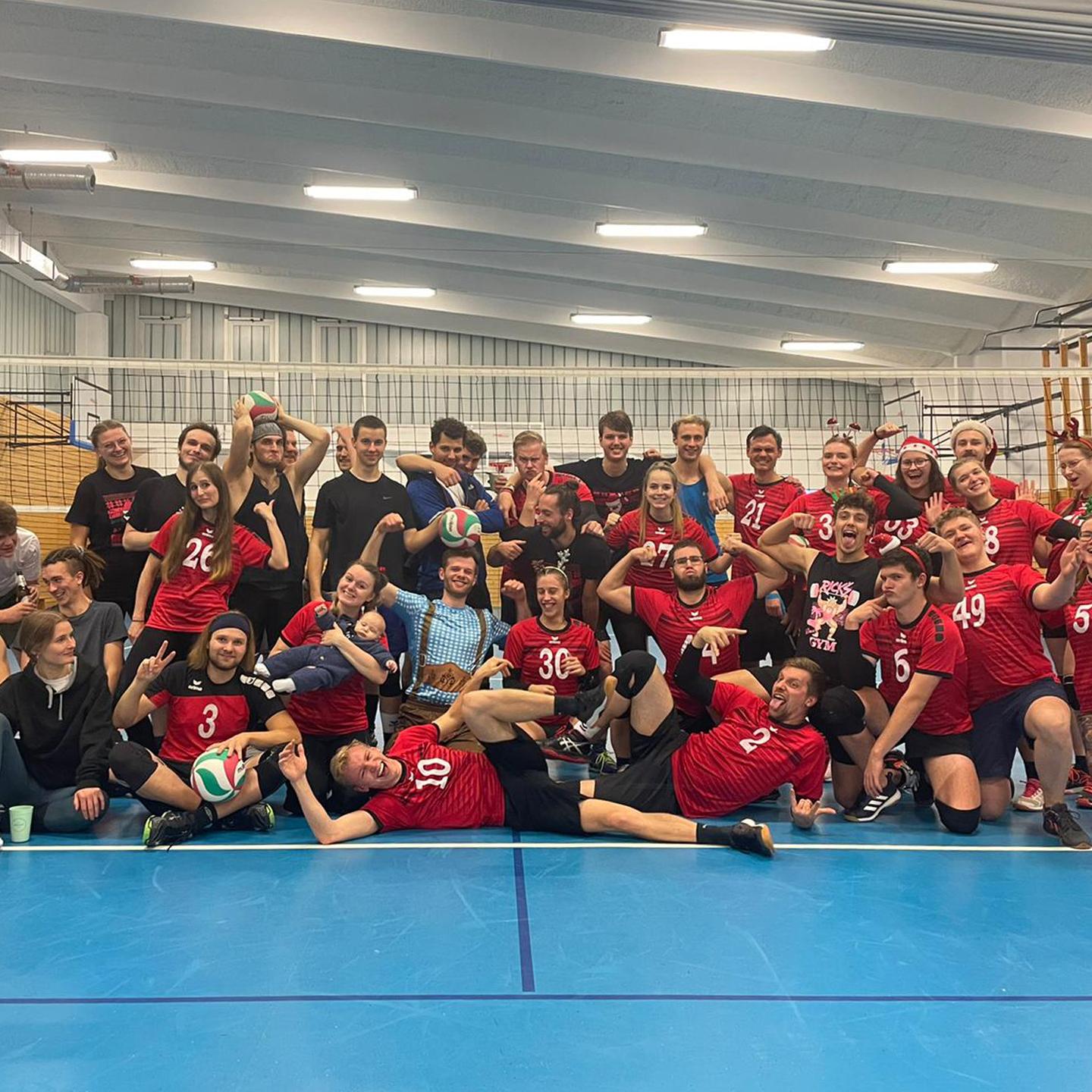 Gruppennaufnahme von Volleyballspielern in roten Trikots, die jubelnd auf einem Trainingsfeld stehen.