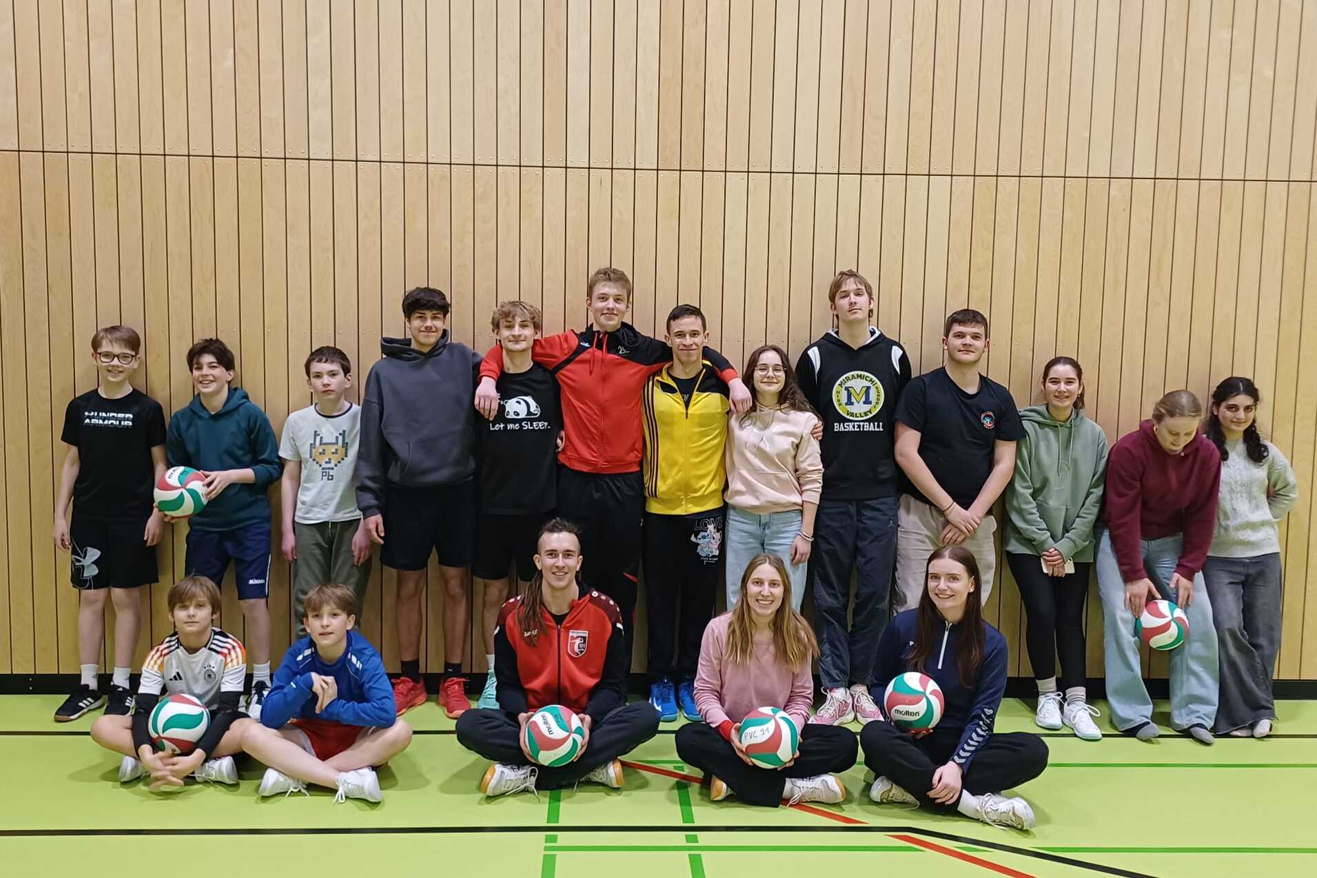Gruppe von Jugendlichen und jungen Erwachsenen mit Volleyballen in einer Sporthalle vor einer Holzpaneelwand.