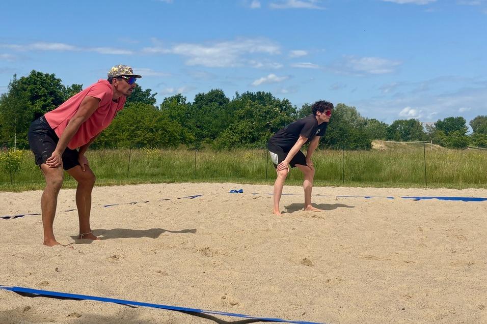 Zwei sportliche Männer in Umkleidung stehen auf einem Sandplatz, bereit für eine Sportaktivität unter blauem Himmel.