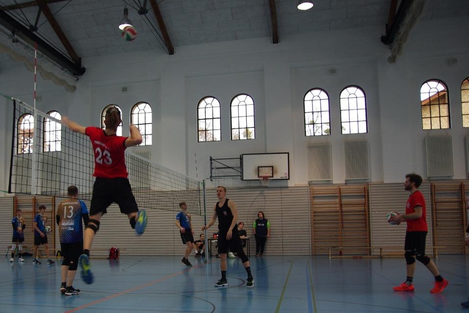 Volleyballspieler springt zum Angriffsschlag in einer Sporthalle mit Fensterfront und Holzbl&ouml;cken.