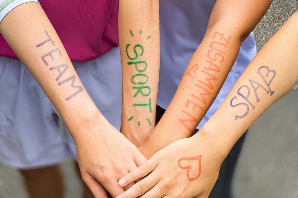 Vier Hände mit bunten Schriftzügen "TEAM", "SPORT", "ZUSAMMEN" und "SPASS" liegen übereinander auf einem grauen Untergrund.
