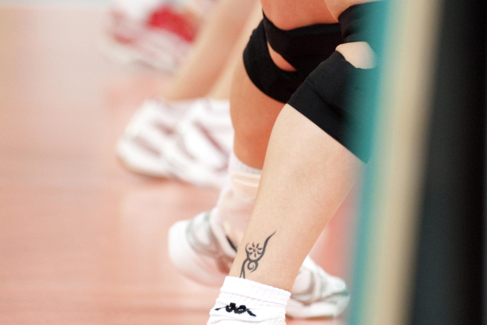 Nahaufnahme der Beine von Volleyballspielerinnen in Sport Schuhen mit einem Tattoo auf der Wade, im Hintergrund der Volleyballplatz.